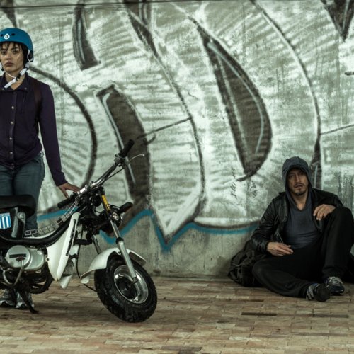 Dos personas, una mujer con casco y moto, un hombre sentado contra una pared.