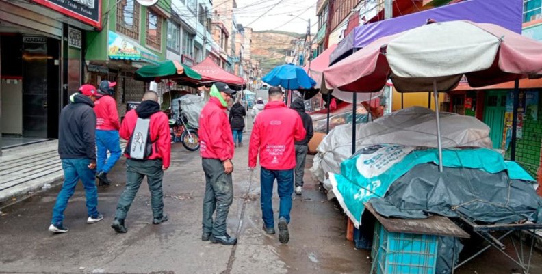 Espacio público en Bogotá Cuatro hombres con chaquetas rojas caminando sobre una calle llena de puestos de venta ambulante