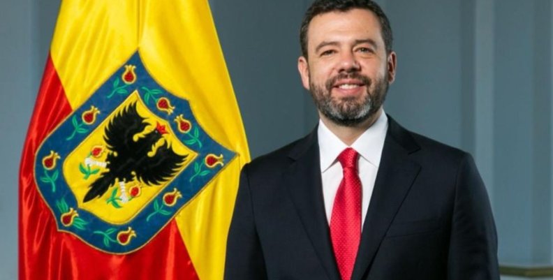 Alcalde Galán Hombre sonriente con traje negro, camisa blanca y corbata roja, delante de un fondo gris, y a su izquierda una bandera roja y amarilla