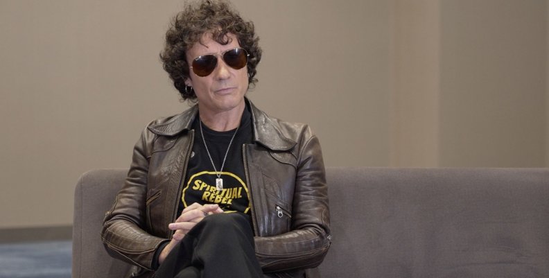 Enrique Bunbury Un hombre con chaqueta de cuero café, pantalón negro y lentes de sol sentado en un sofá