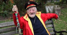 Un hombre anciano vistiendo una chaqueta roja, un bastón y un sombrero militar