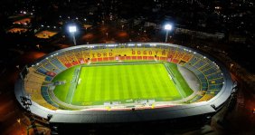Vista panorámica de un estadio. Las gradas son amarillas con rojo y hay una chancha de fútbol en el medio