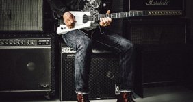 Una persona sentada, tocando una guitarra eléctrica blanca. Viste chaqueta oscura, camiseta gráfica, jeans y botas de cuero. A su alrededor hay varios amplificadores de guitarra