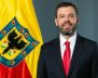 Hombre sonriente con traje negro, camisa blanca y corbata roja, delante de un fondo gris, y a su izquierda una bandera roja y amarilla