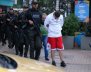 Policías armados custodiando a un hombre con hoodie blanco y shorts rojos