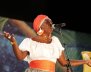Una mujer con un turbante naranja y una camisa blanca canta frente a un micrófono.