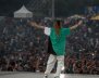 Hombre en el escenario con camisa verde y pantalón blanco abre los brazos frente al público