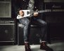 Una persona sentada, tocando una guitarra eléctrica blanca. Viste chaqueta oscura, camiseta gráfica, jeans y botas de cuero. A su alrededor hay varios amplificadores de guitarra