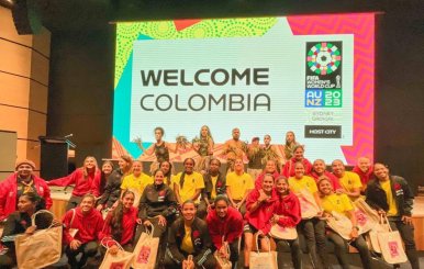 Mujeres de la Selección Colombia femenina, usando chaquetas amarillas y rojas, posan reunidas frente a un letrero que dice 'Bienvenido Colombia'