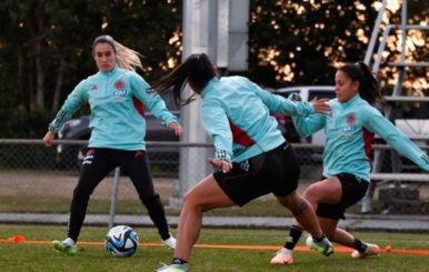 Tres mujeres con uniforme color celeste disputando un balón blanco, paradas en pasto color verde