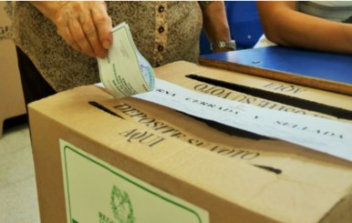 Una caja que funge como urna de votación con una ranura encima por la cual una mano va a introducir un papel de voto.