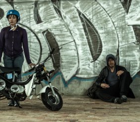 Dos personas, una mujer con casco y moto, un hombre sentado contra una pared.