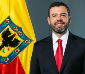 Hombre sonriente con traje negro, camisa blanca y corbata roja, delante de un fondo gris, y a su izquierda una bandera roja y amarilla