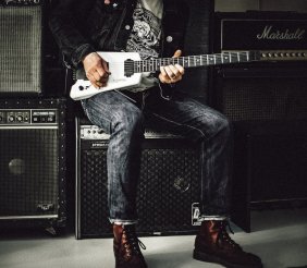 Una persona sentada, tocando una guitarra eléctrica blanca. Viste chaqueta oscura, camiseta gráfica, jeans y botas de cuero. A su alrededor hay varios amplificadores de guitarra