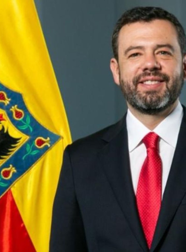 Hombre sonriente con traje negro, camisa blanca y corbata roja, delante de un fondo gris, y a su izquierda una bandera roja y amarilla